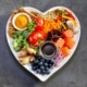 Heart healthy foods, good for lowering cholesterol, in a heart-shaped dish including acai, lentils, soy sauce, ginger, salmon, carrot, tomato, turmeric, cinnamon, walnuts, garlic, peppers, broccoli, basil, onion