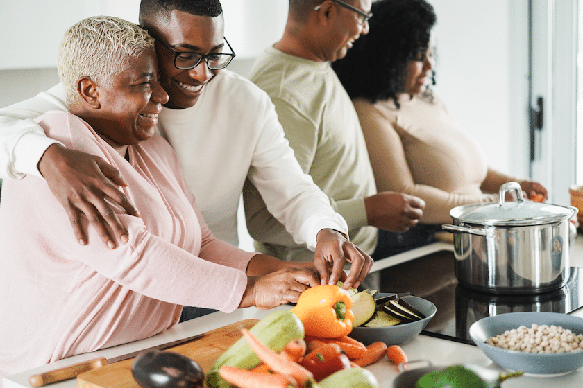 Family cooking a meal together, older woman and young man side-hug and smile fondly while preparing vegetables, other family members cook at the stove in the background