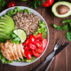 Bowl with leafy greens, quinoa, chicken, tomato, and lime wedges sit on top of a wooden table with silverware.